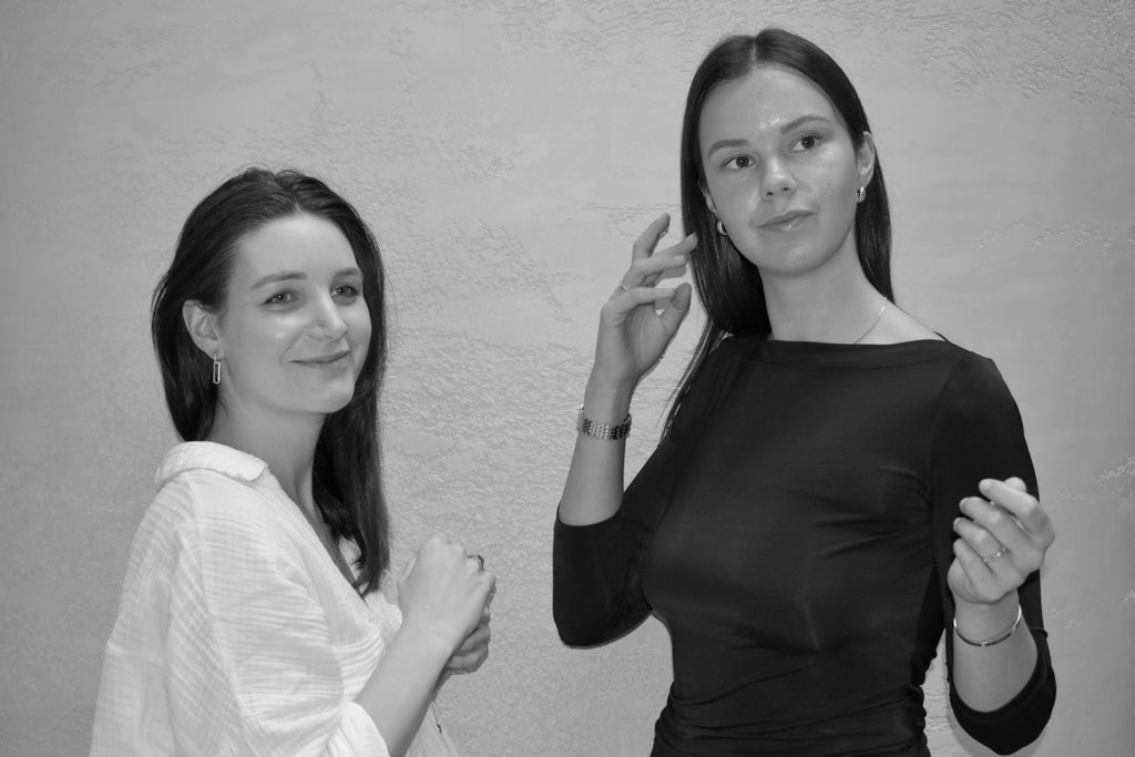 Two women stand together against a textured wall. One smiles softly at the camera, wearing a light blouse, while the other, in a dark top, looks to the side and adjusts her hair. The photo is in black and white.