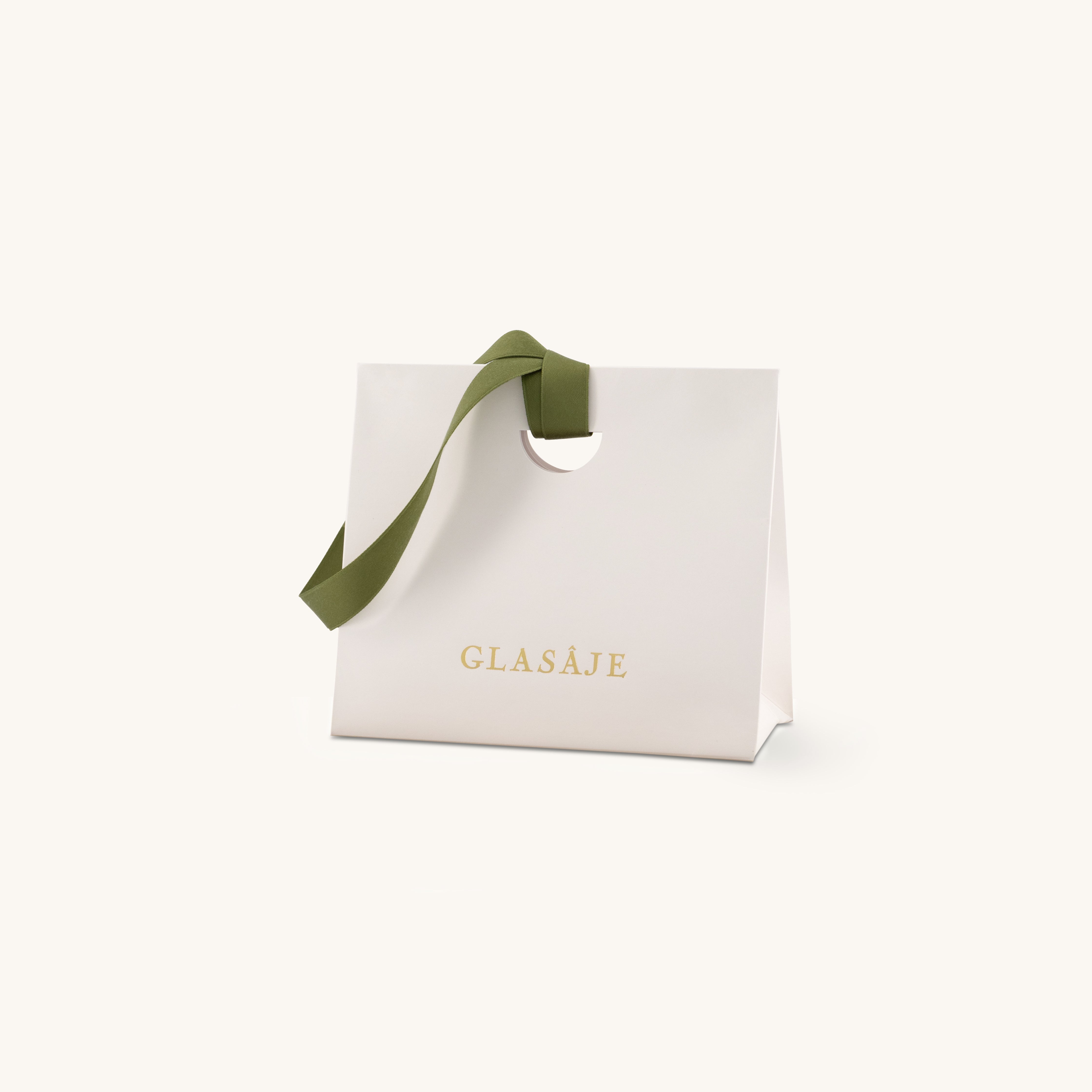 A white gift bag with a cut-out handle, olive green ribbon, and the word GLASÁJE in gold letters on the front, set against a plain light background.