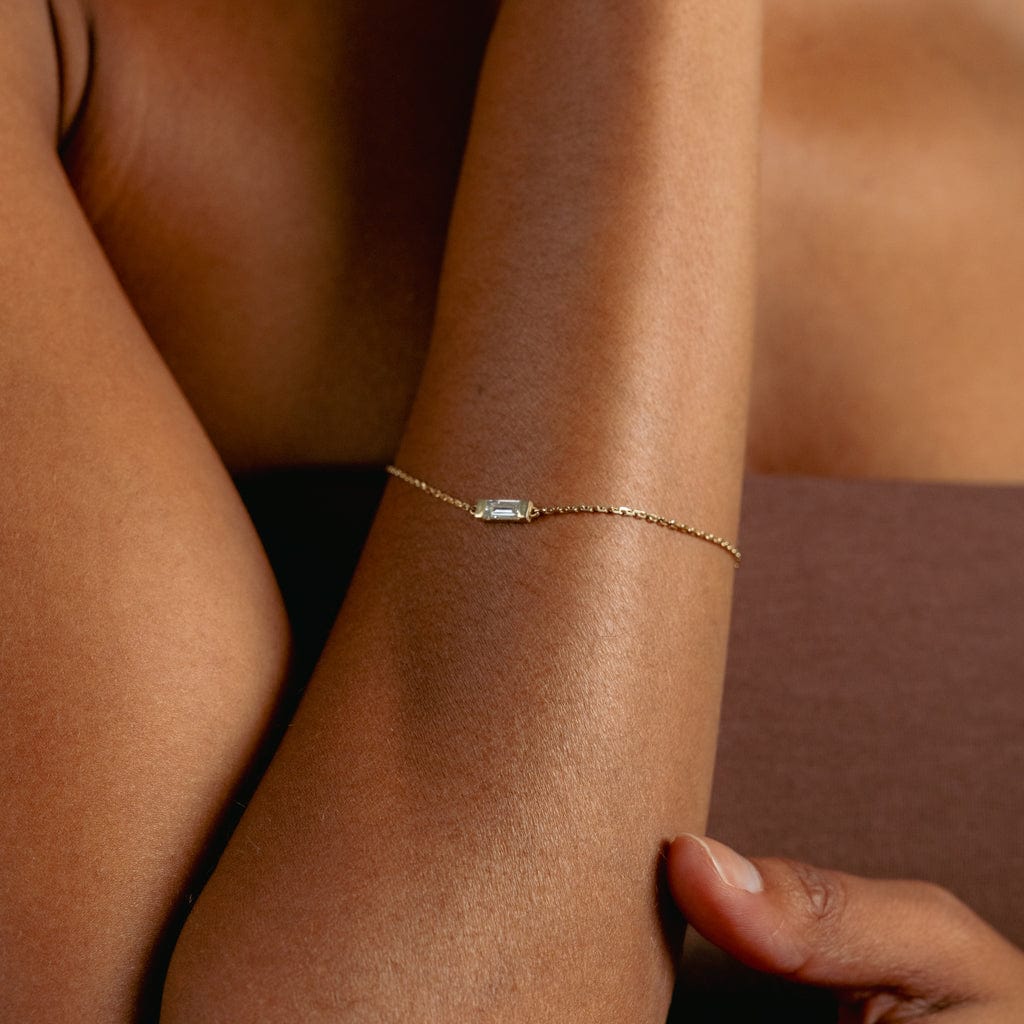 A close-up of a persons arm wearing a delicate gold chain bracelet with a small rectangular gemstone, against a brown background. The persons skin is medium brown and their hand rests gently on their arm.