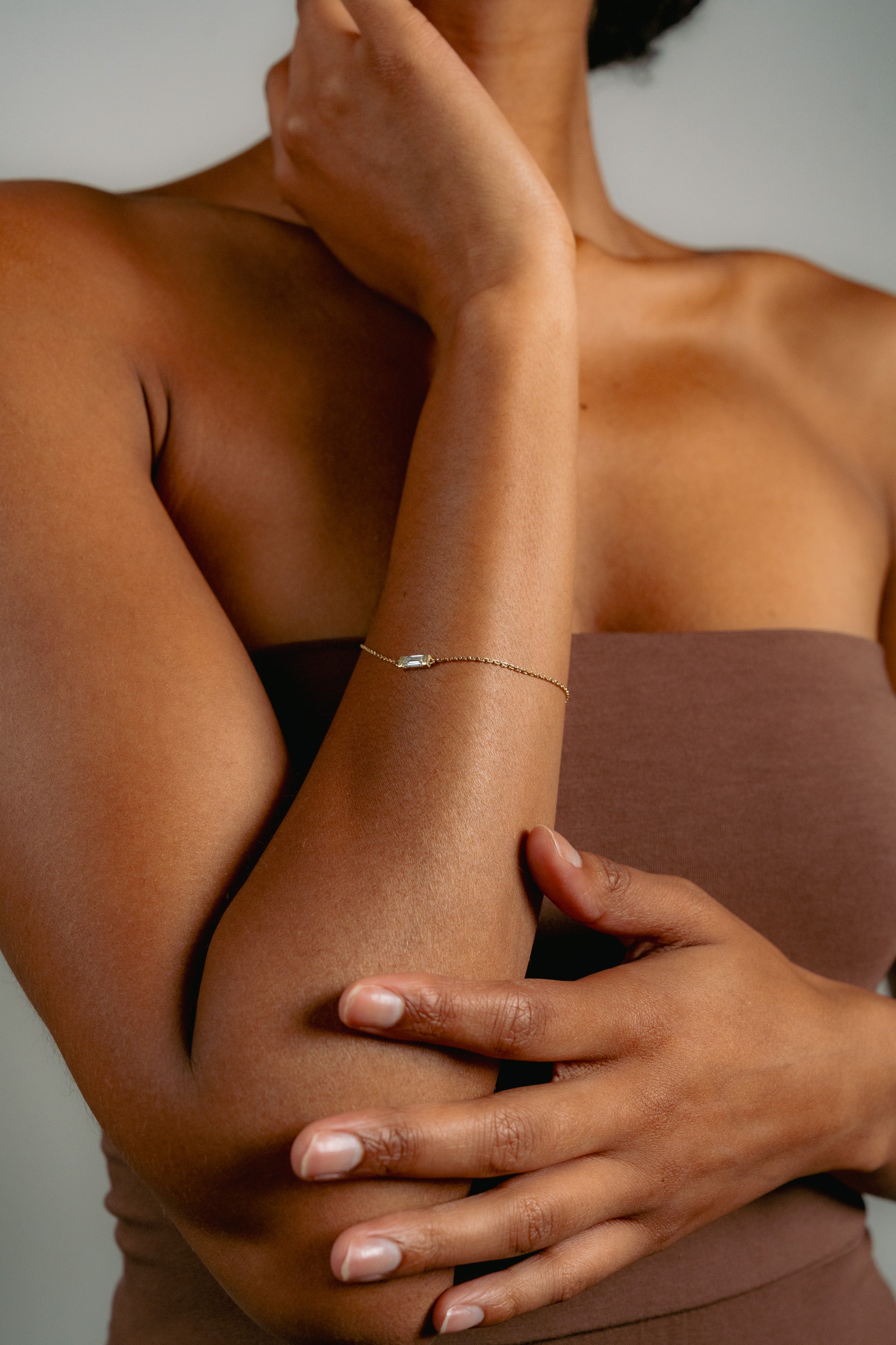 A woman wearing a brown strapless top poses with one arm crossed over her body and the other hand touching her neck, showcasing a delicate gold bracelet with a small charm.
