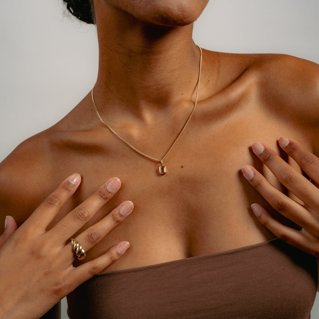A woman wearing a brown strapless top shows off a delicate gold necklace with a small pendant and a gold twisted ring. Her hands are gently touching her collarbone, and the background is neutral.