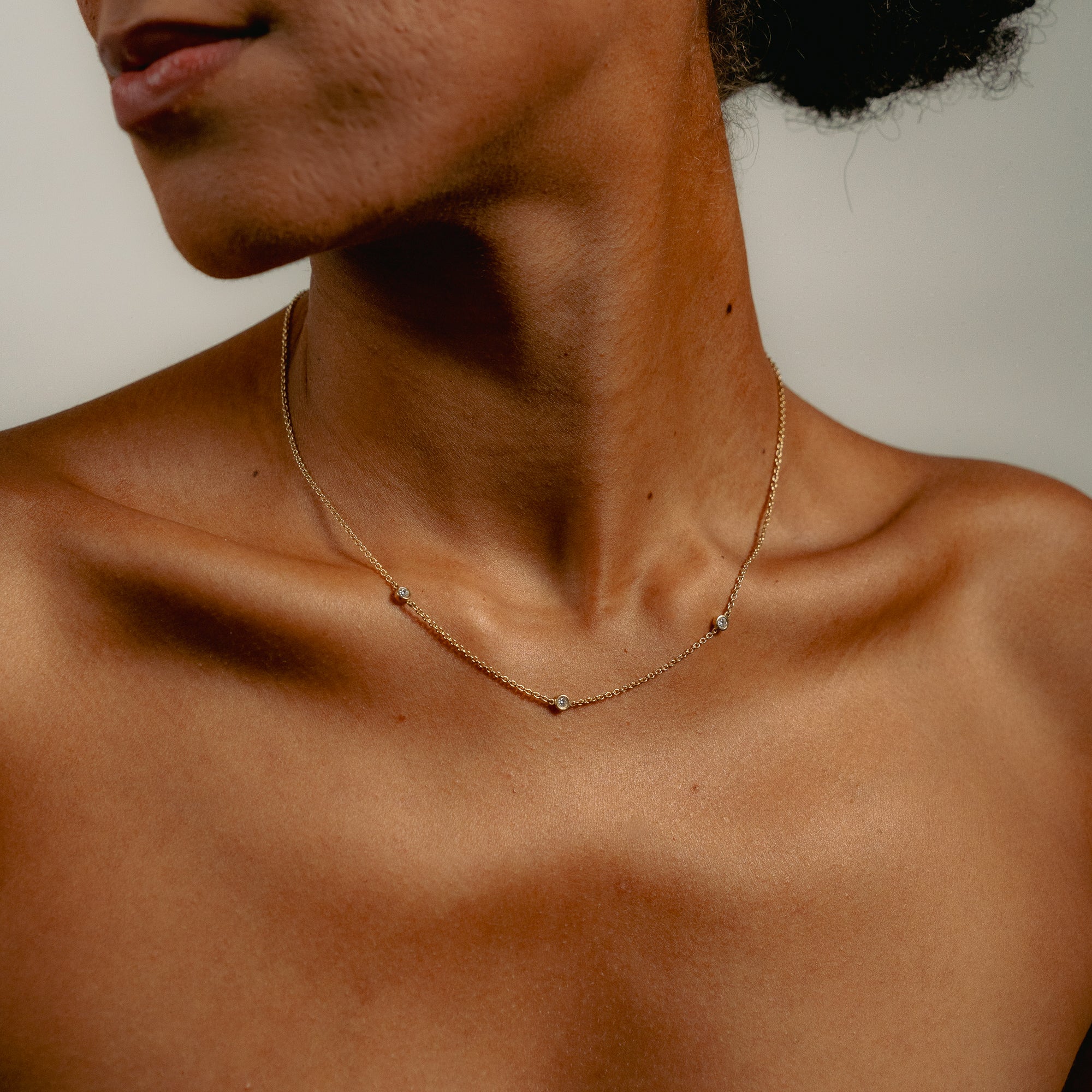 A close-up of a woman's neck and collarbone wearing a delicate gold necklace with 3 three round diamonds against a plain background.