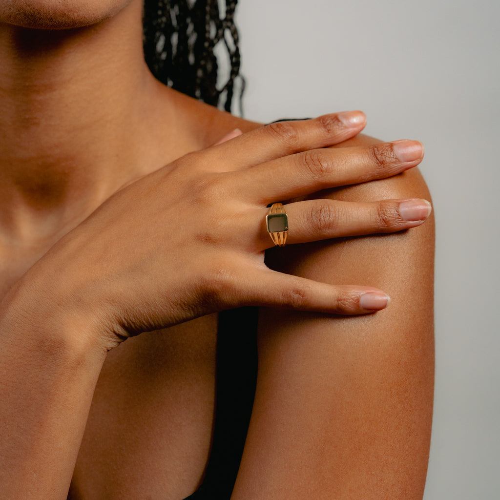 A close-up of a person’s hand resting on their shoulder, showing a gold ring with a rectangular design on their finger. The background is plain and the person is wearing a black top.
