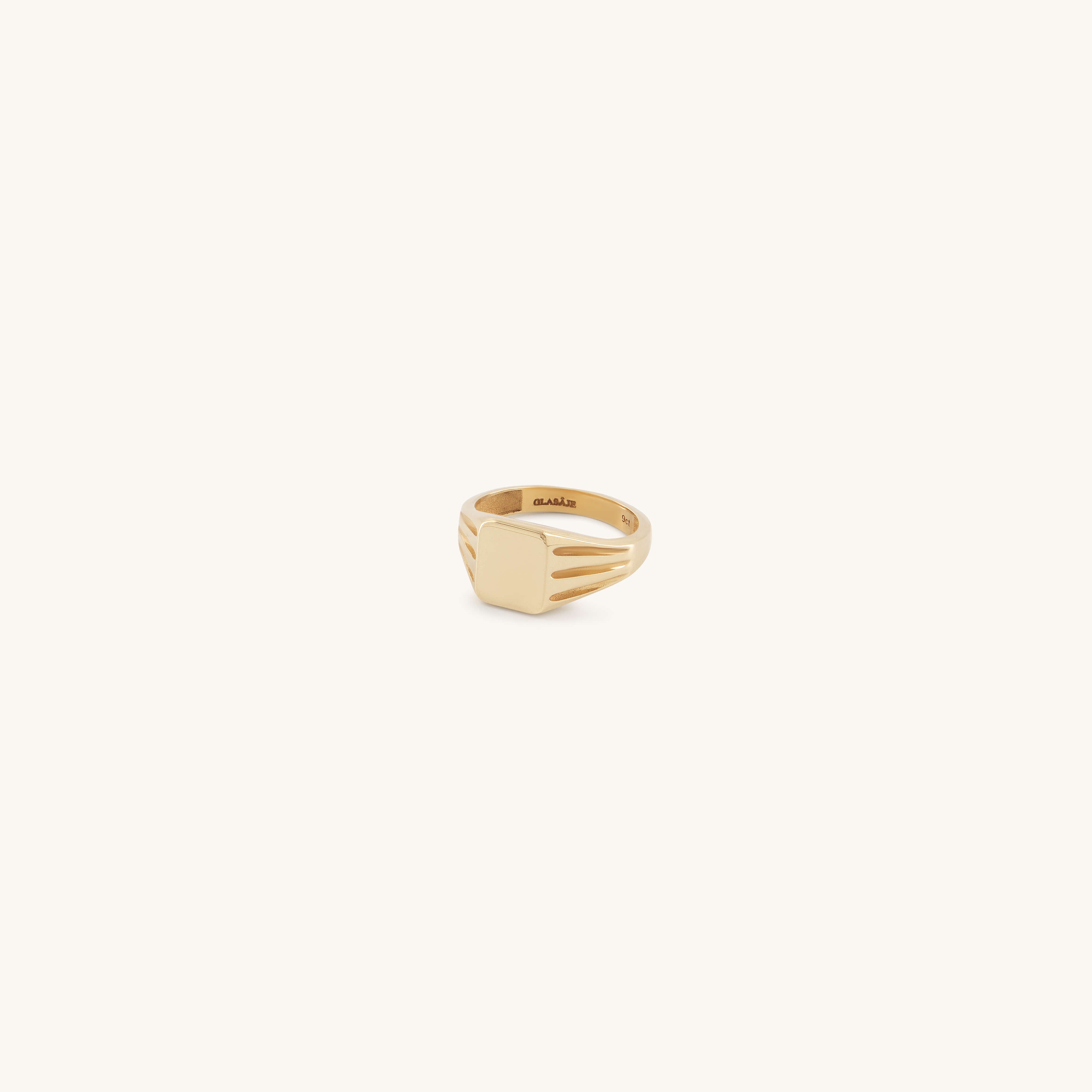 The Glasâje Signet Ring is crafted in solid gold with a rectangular flat face and debossed details, plus decorative grooves on the band, showcased against a light cream background.