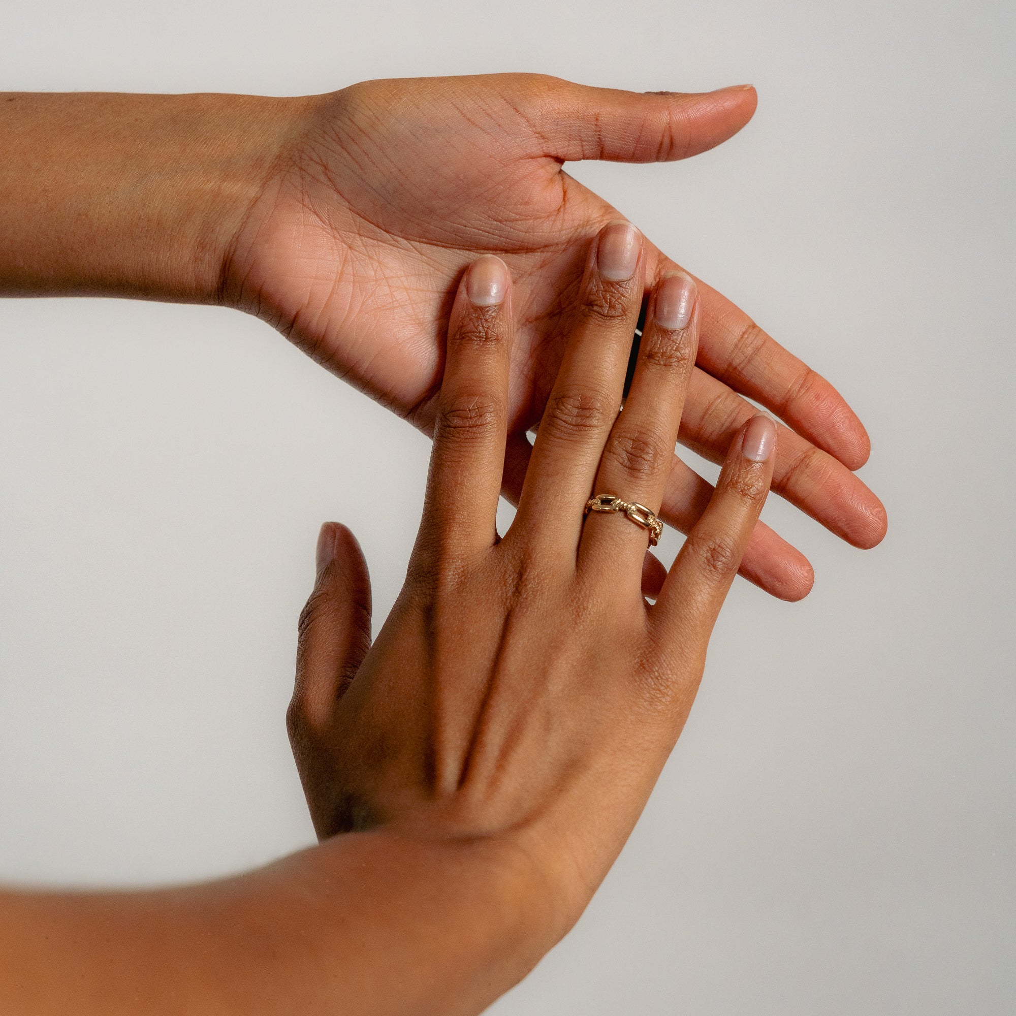 Two hands with warm skin tones interact gently. One hand wears a gold chain ring, conveying connection and tenderness against a soft gray background.