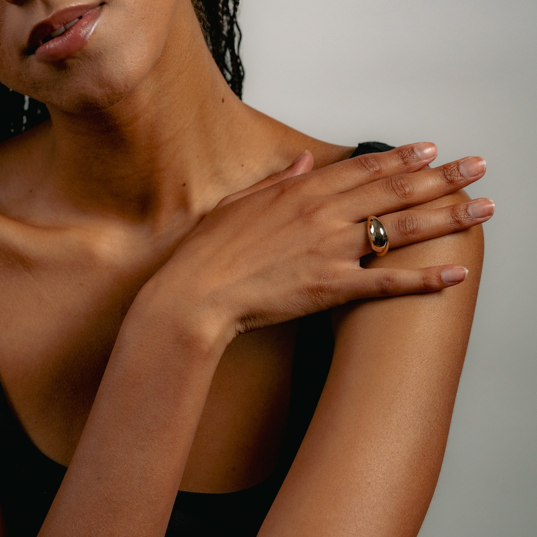 A person with braided hair rests a hand on their shoulder, wearing the solid gold Glasâje Dôme Ring. A black top and neutral background highlight the lower face and upper body, drawing attention to the ring.