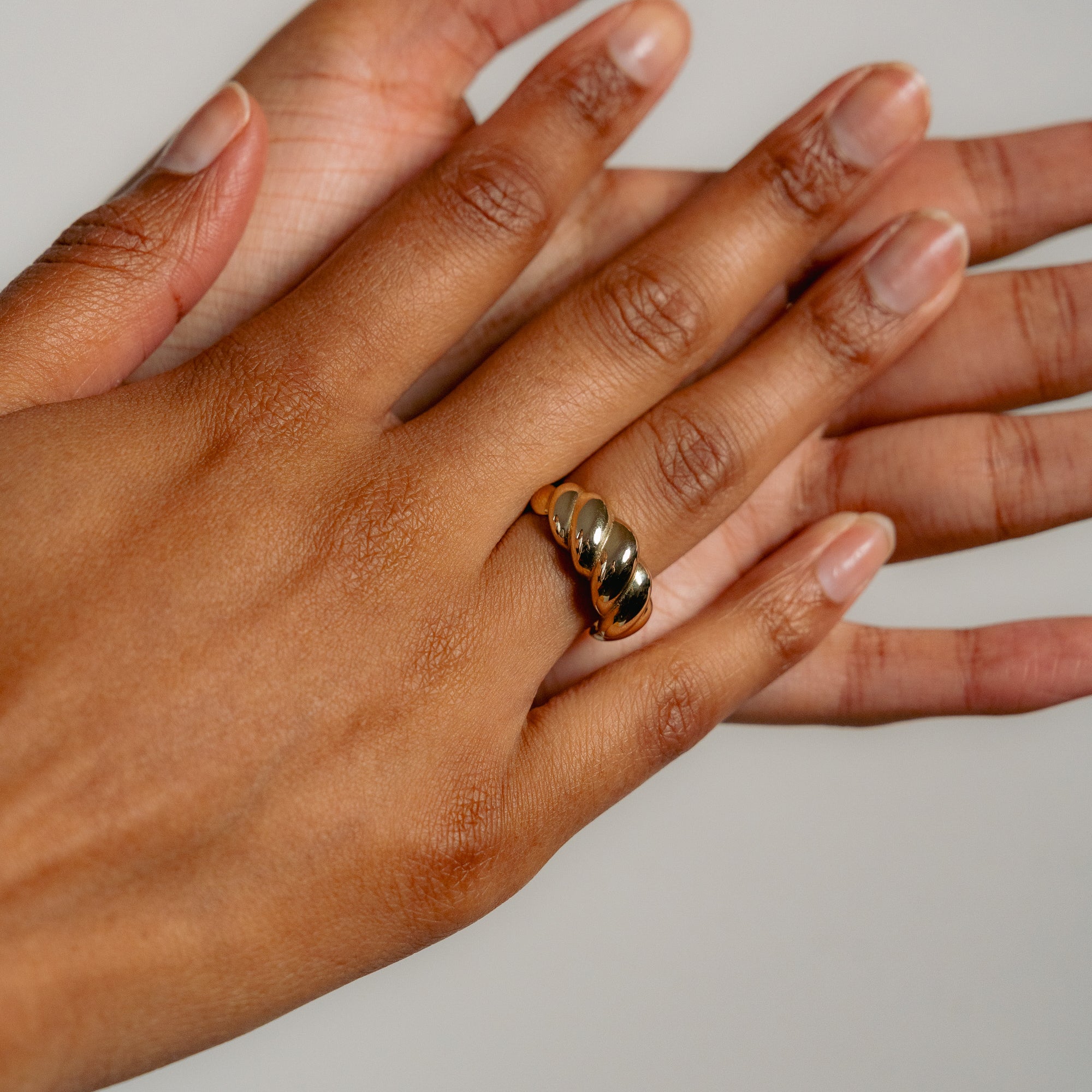 A close-up shows a hand wearing Glasâje’s shiny, chunky Lune Ring with a croissant-inspired design, while another hand gently holds it from below. The nails are natural and well-groomed against a plain, neutral background.