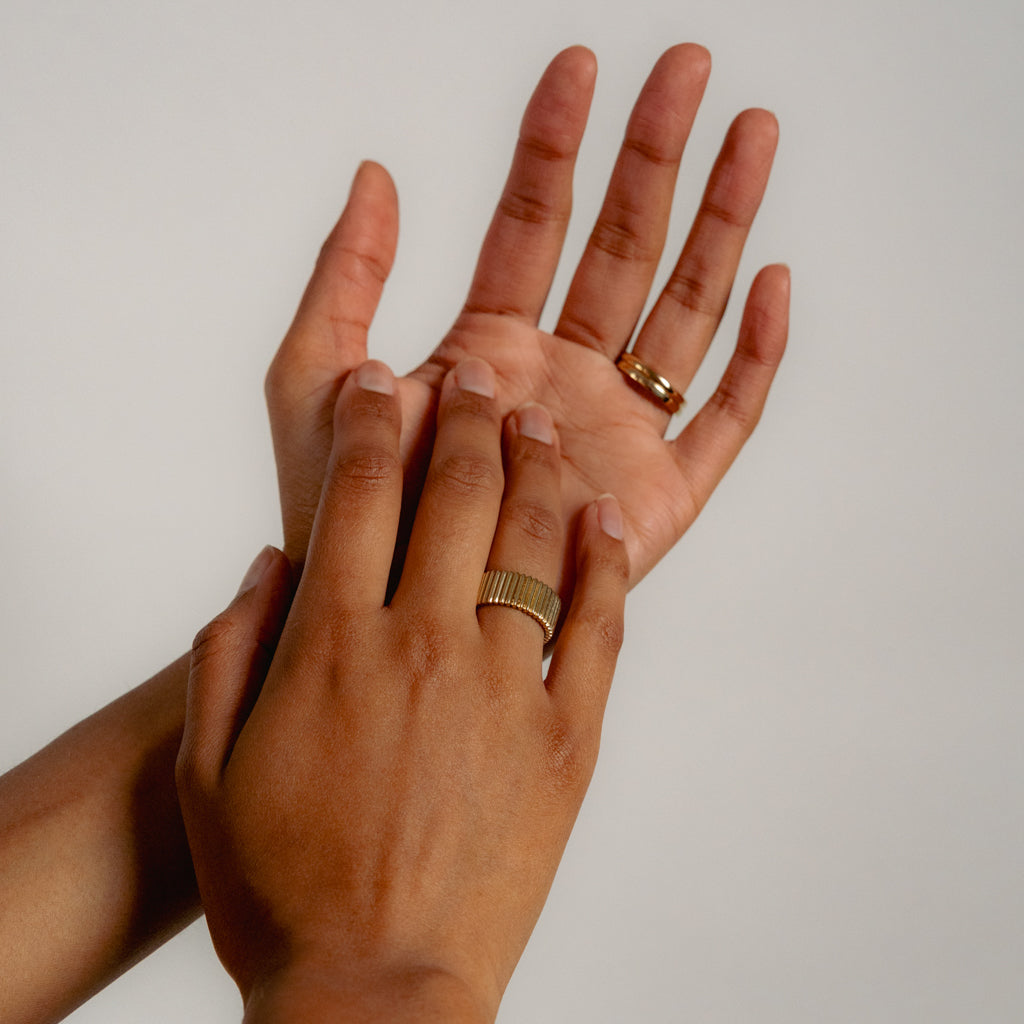 Two hands with brown skin tones are shown against a plain background. One hand wears a thick gold ring on the middle finger, while the other hand wears a thin gold ring on the ring finger.