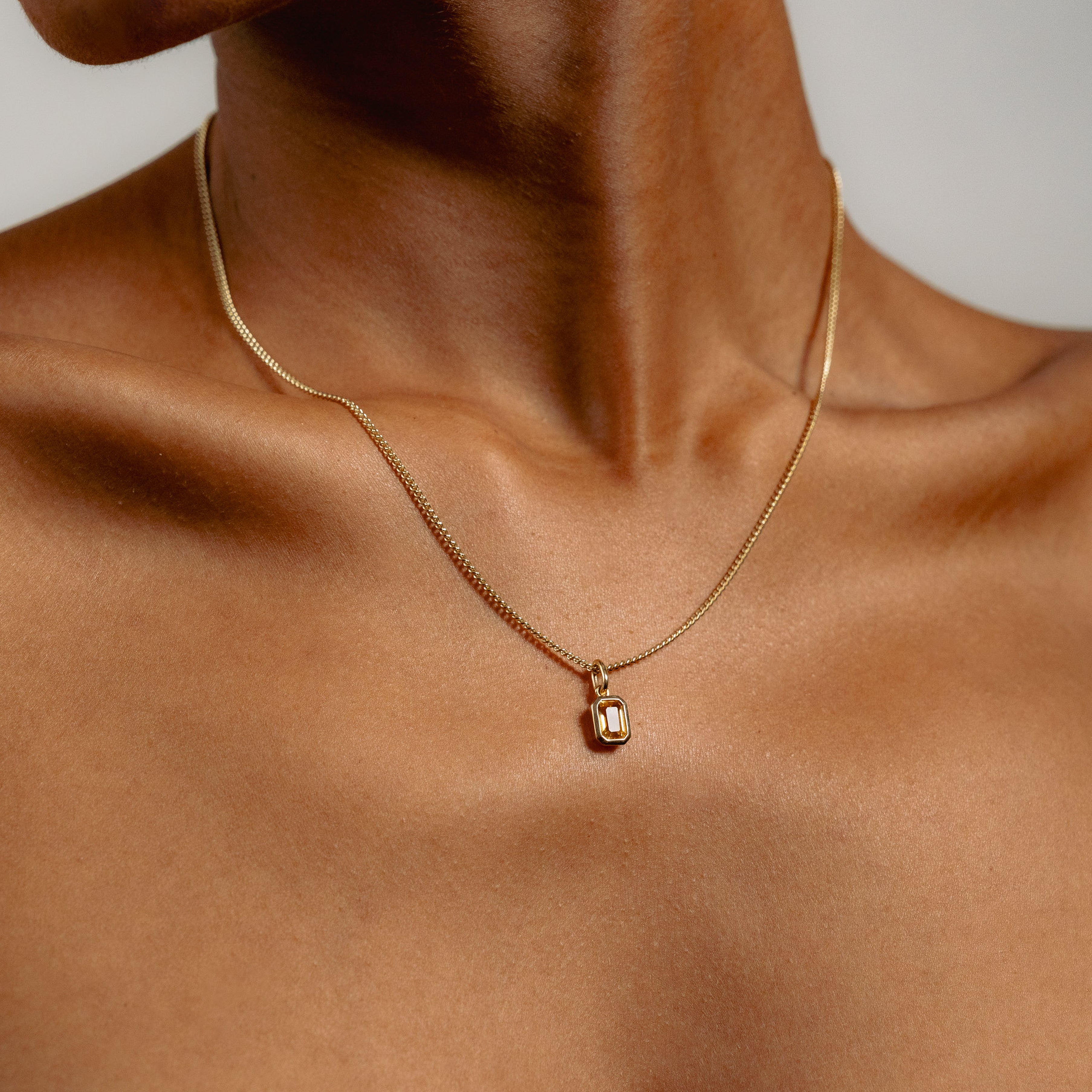 Close-up of a person wearing a delicate gold necklace with a rectangular auburn citrine pendant. The skin is smooth, and the image exudes a sense of elegance and simplicity.