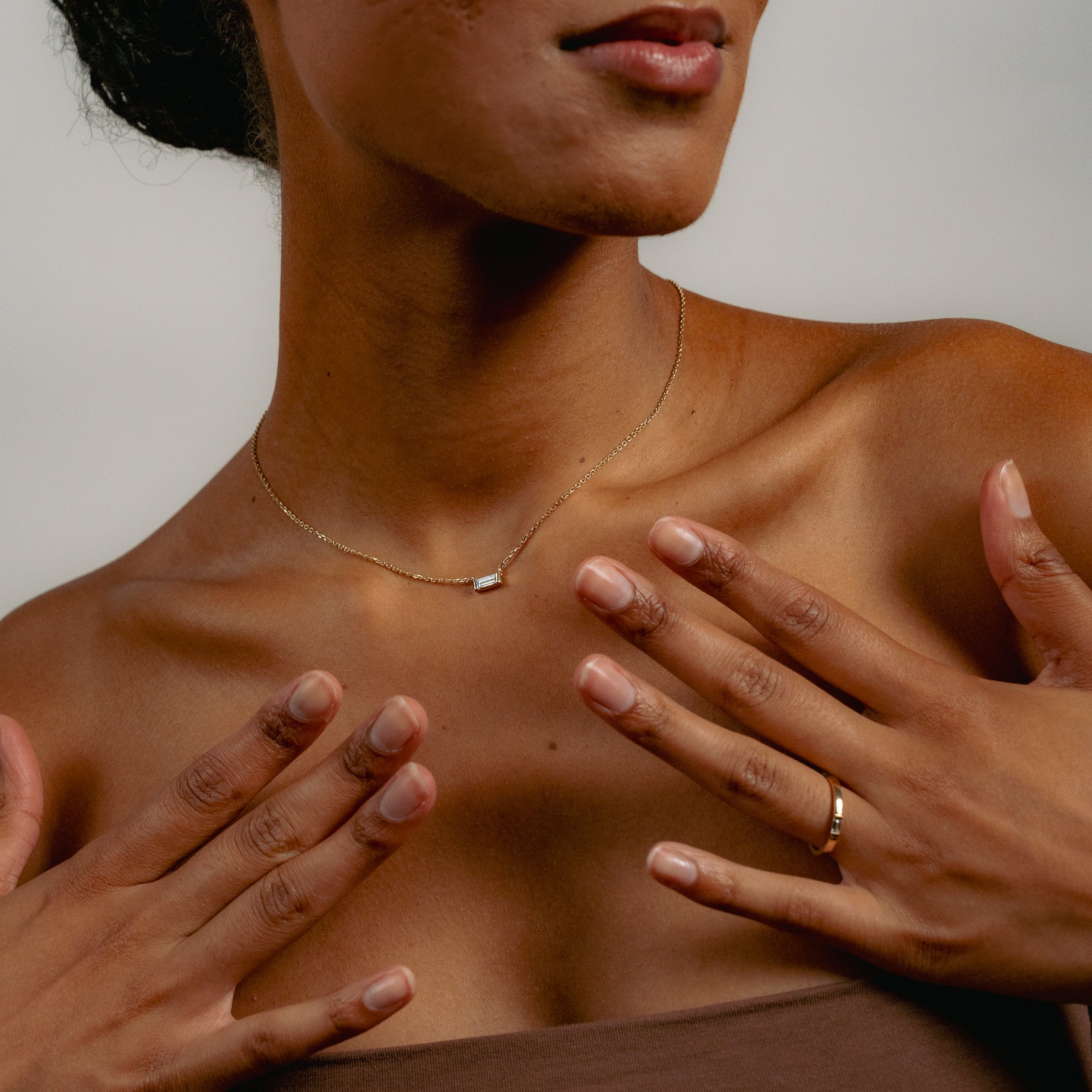 A woman wearing a strapless brown top touches her collarbone with both hands, displaying a gold necklace with a small baguette diamond pendant and a stacking diamond ring on her finger against a neutral background.