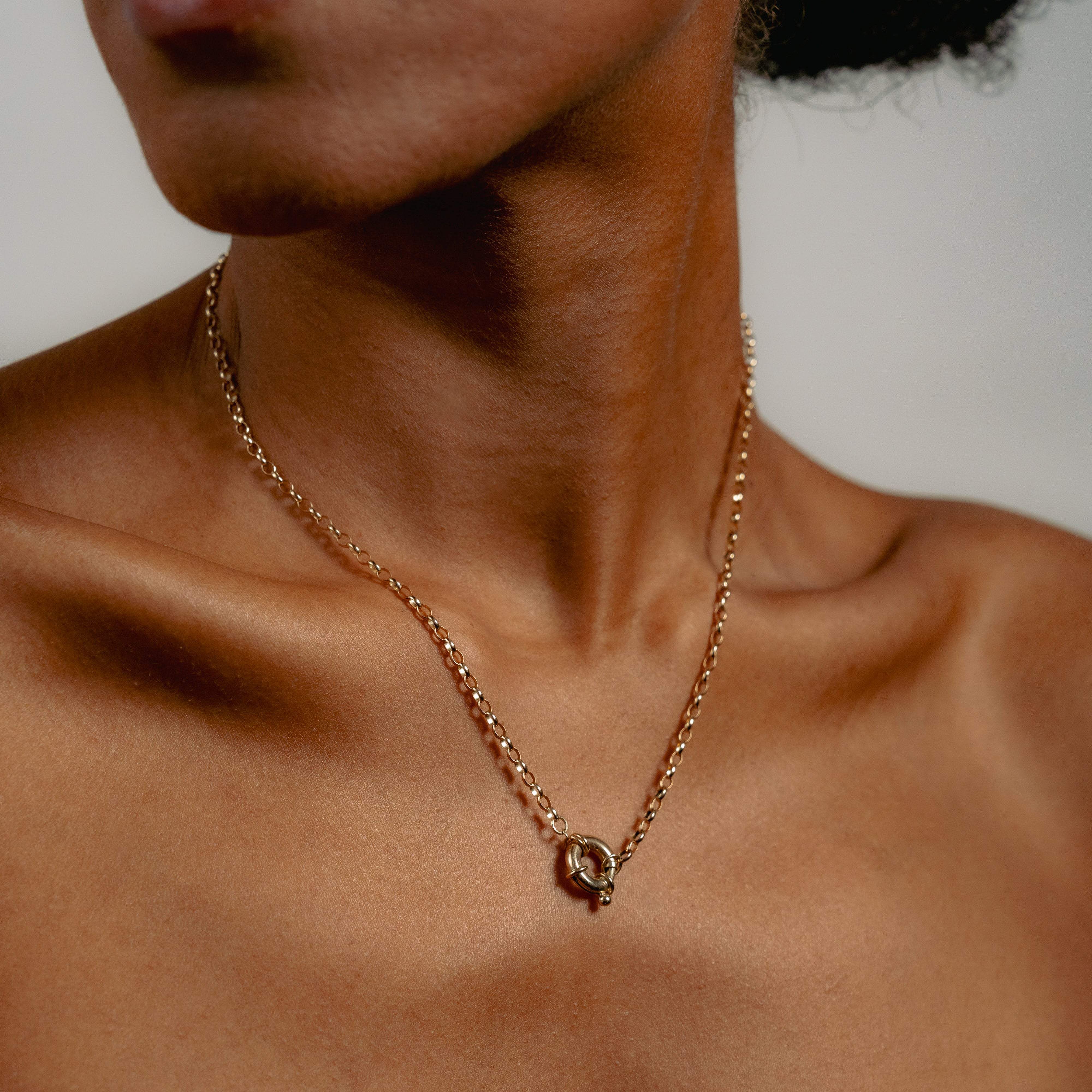 A close-up shows a person’s neck and collarbone wearing the Glasâje Hosier Necklace, which features a gold belcher chain with a circular clasp at the center. The background is neutral and blurred.