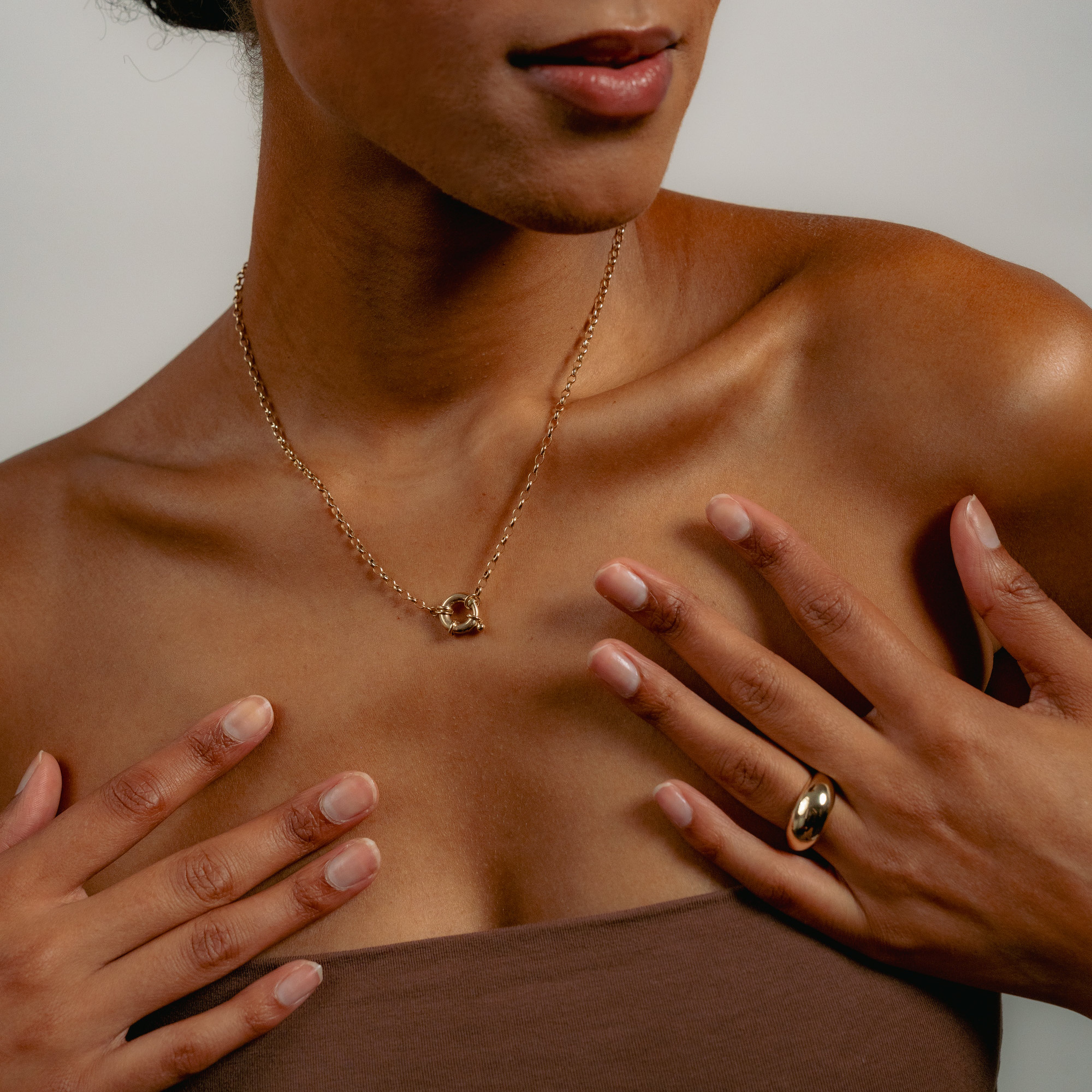 A woman in a strapless brown top showcases the Glasâje Hosier Necklace with a belcher chain and matching gold ring, her manicured hands resting on her collarbone against a neutral background.