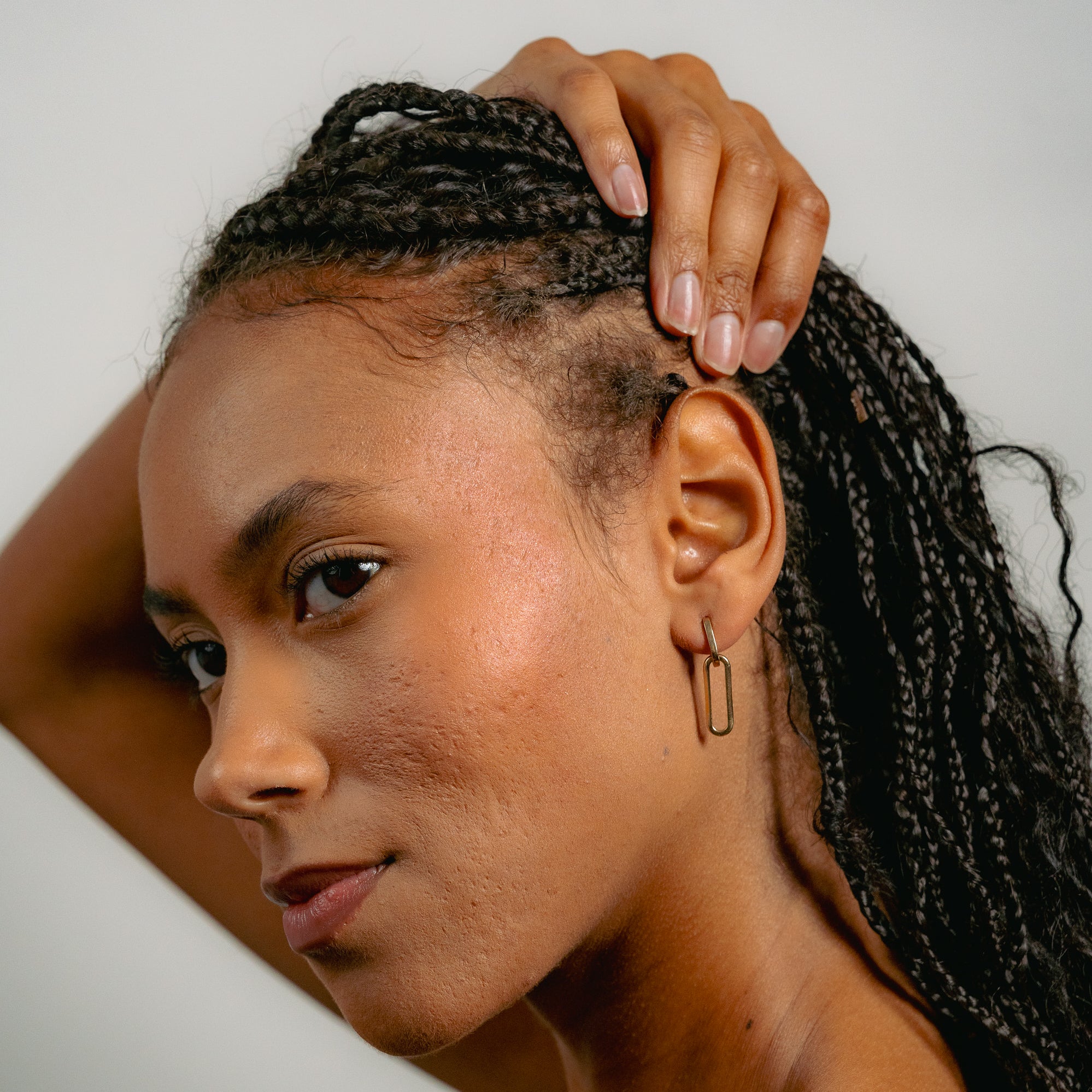 A woman with braided hair touches her head, revealing her scalp. She confidently showcases Glasâje Paperclip Earrings, highlighting their versatile style against a plain background.