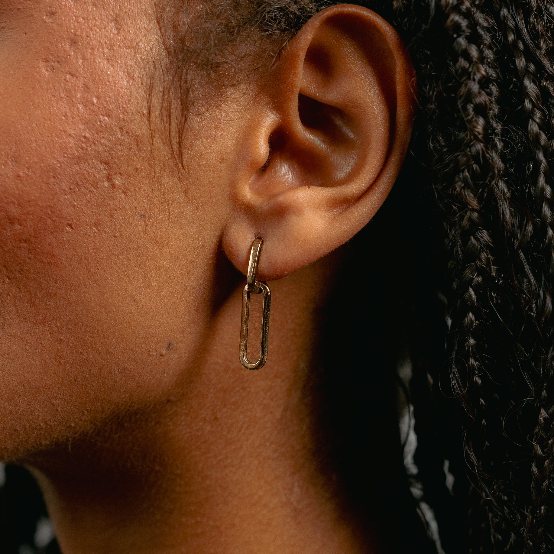 Close-up of a person with medium brown skin and braided hair wearing Glasâje’s 9ct solid gold Paperclip Earrings—rectangular studs ideal for versatile styling.