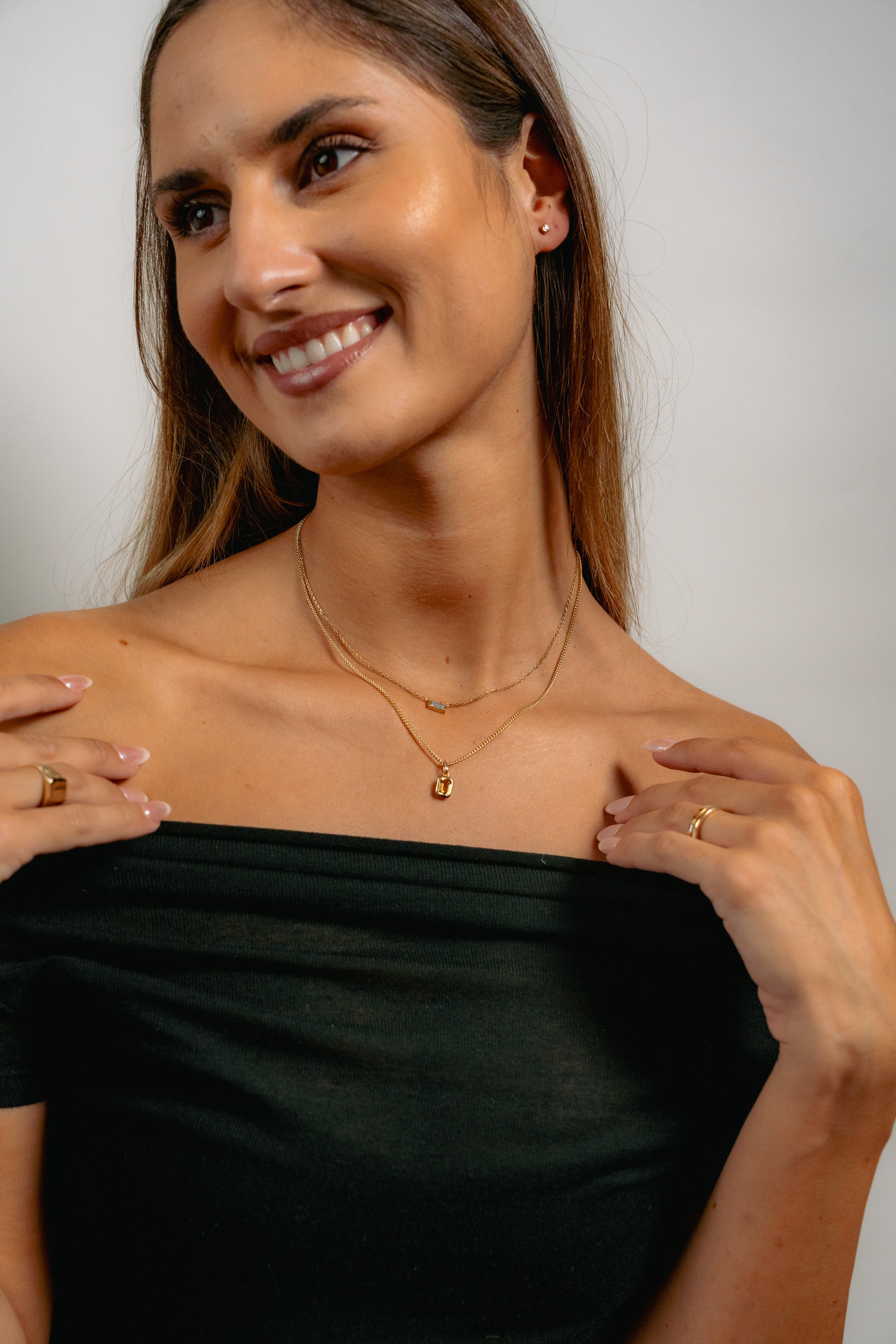 A smiling woman with long brown hair wears a black off-the-shoulder top, gold rings, and layered gold necklaces. She is looking to the side against a plain background.
