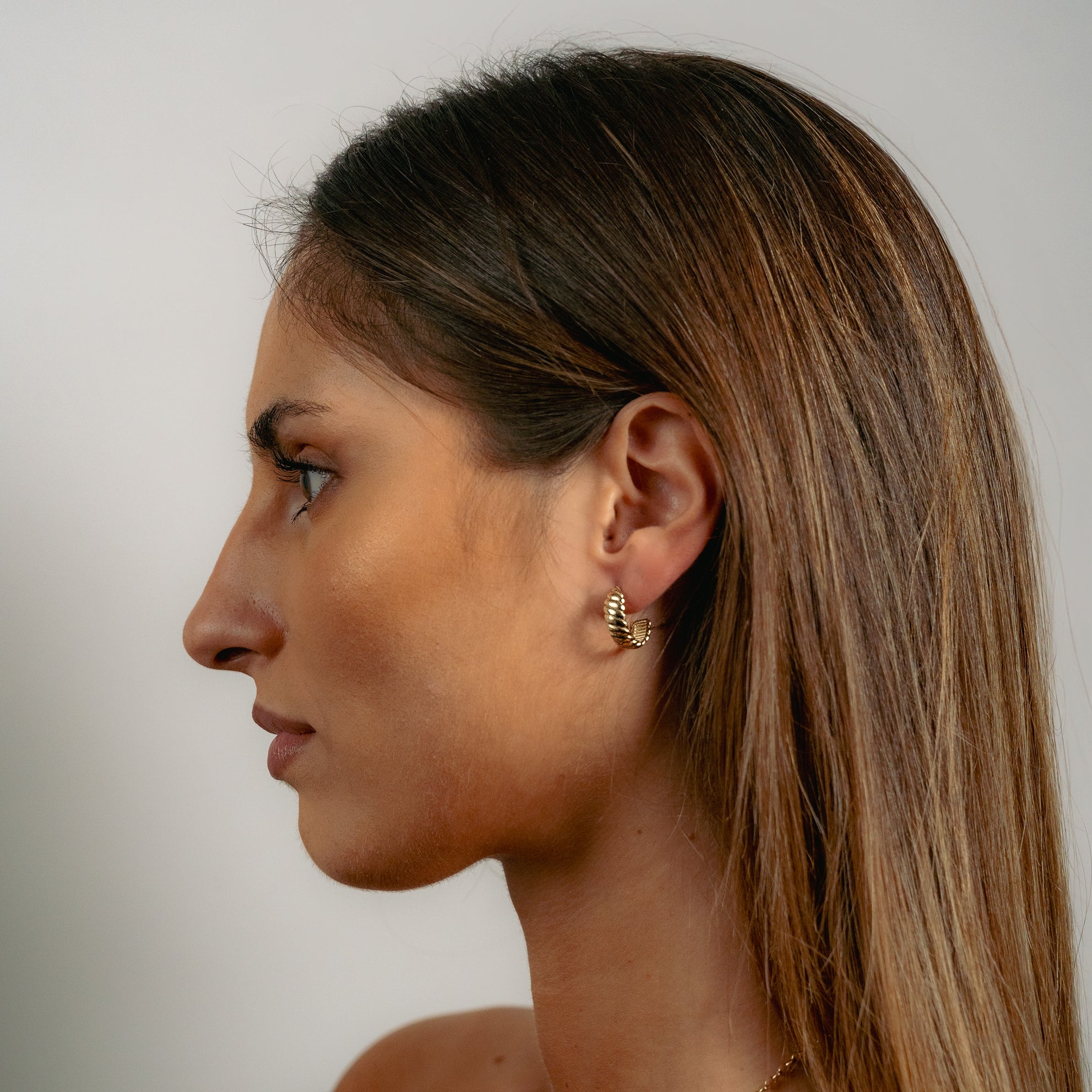 Profile view of a woman with long brown hair wearing a gold twisted hoop earring. The background is a soft white.