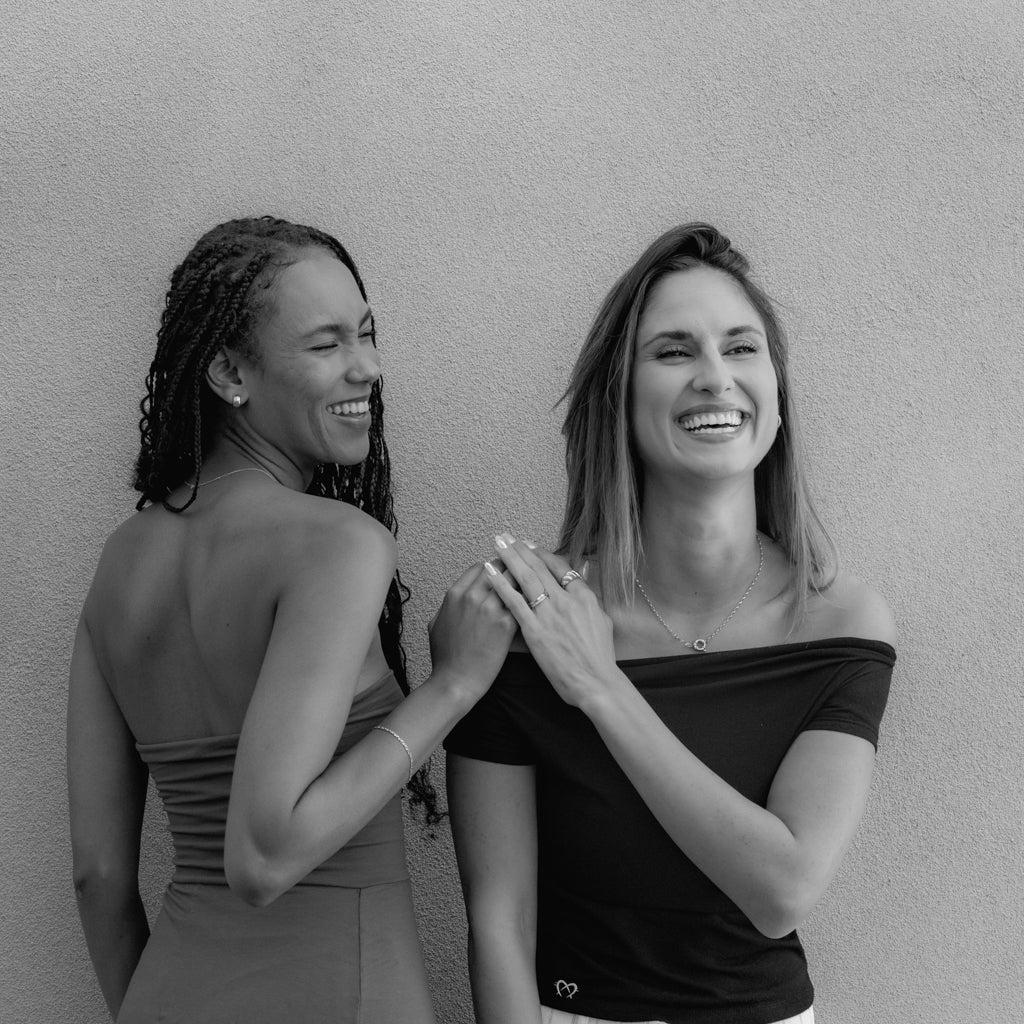 Two women smiling and laughing together in front of a plain wall. One has braided hair and wears a strapless top, while the other has straight hair and wears an off-the-shoulder top. The mood is joyful and friendly.