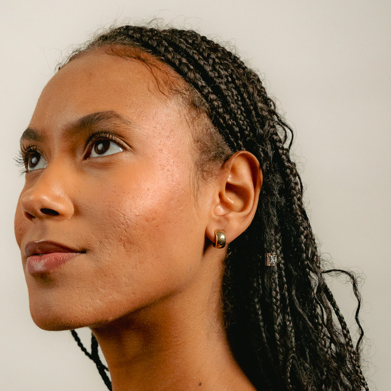 A woman with braided hair looks upward and left, wearing Glasâjes Gabriel Huggies—small solid gold earrings. A plain, light background highlights this timeless staple.
