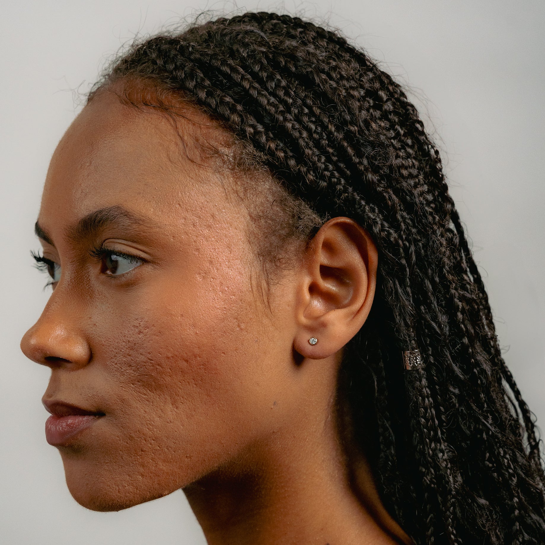 A woman with braided hair and smooth, dark skin is shown in profile, facing left. She wears Glasâjes Satellite Studs in solid gold and has a calm expression against a plain, light background.