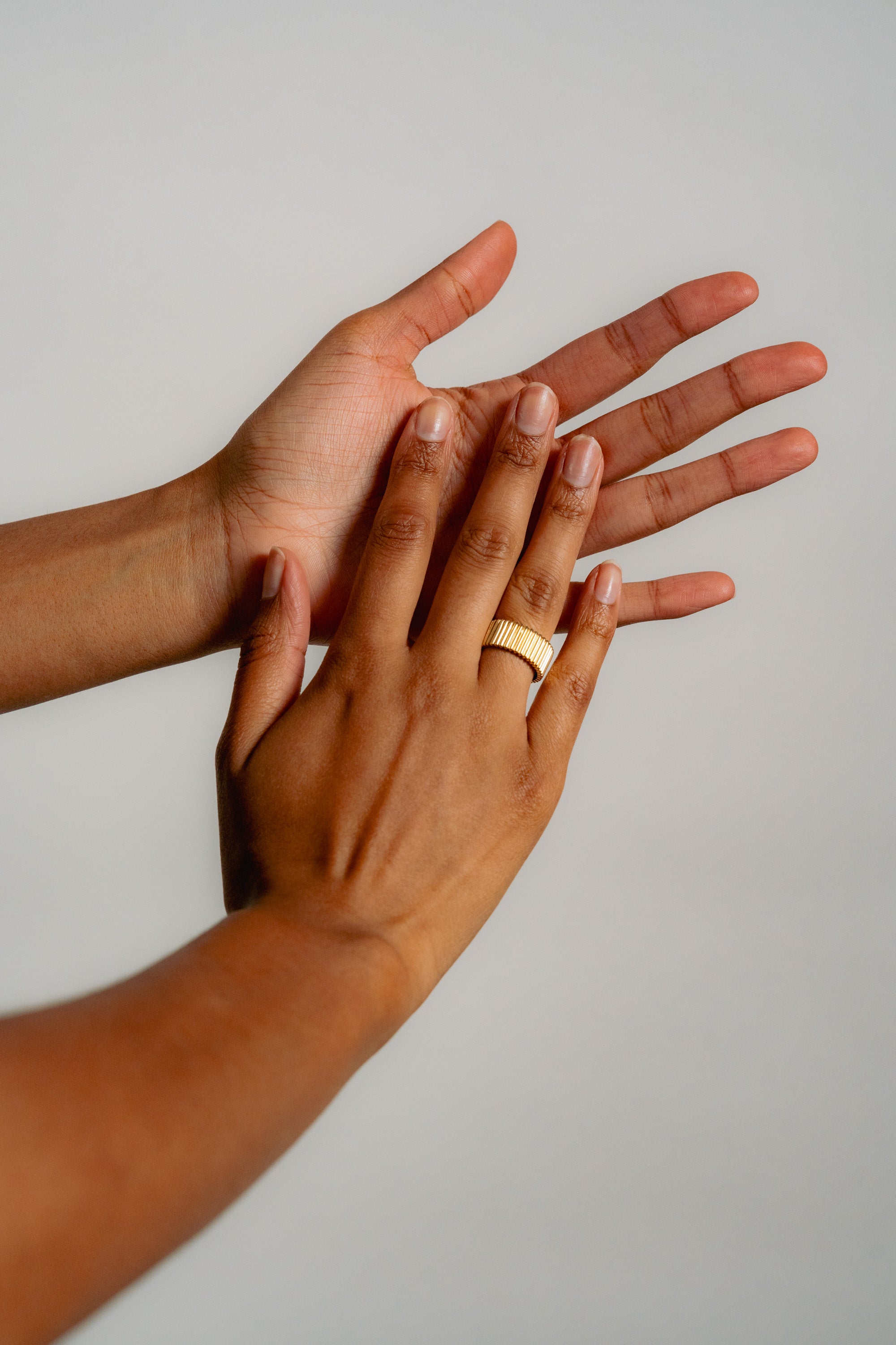 A close-up of two hands with medium brown skin, one resting gently on top of the other. The top hand wears a gold ring. The background is plain and light-colored.