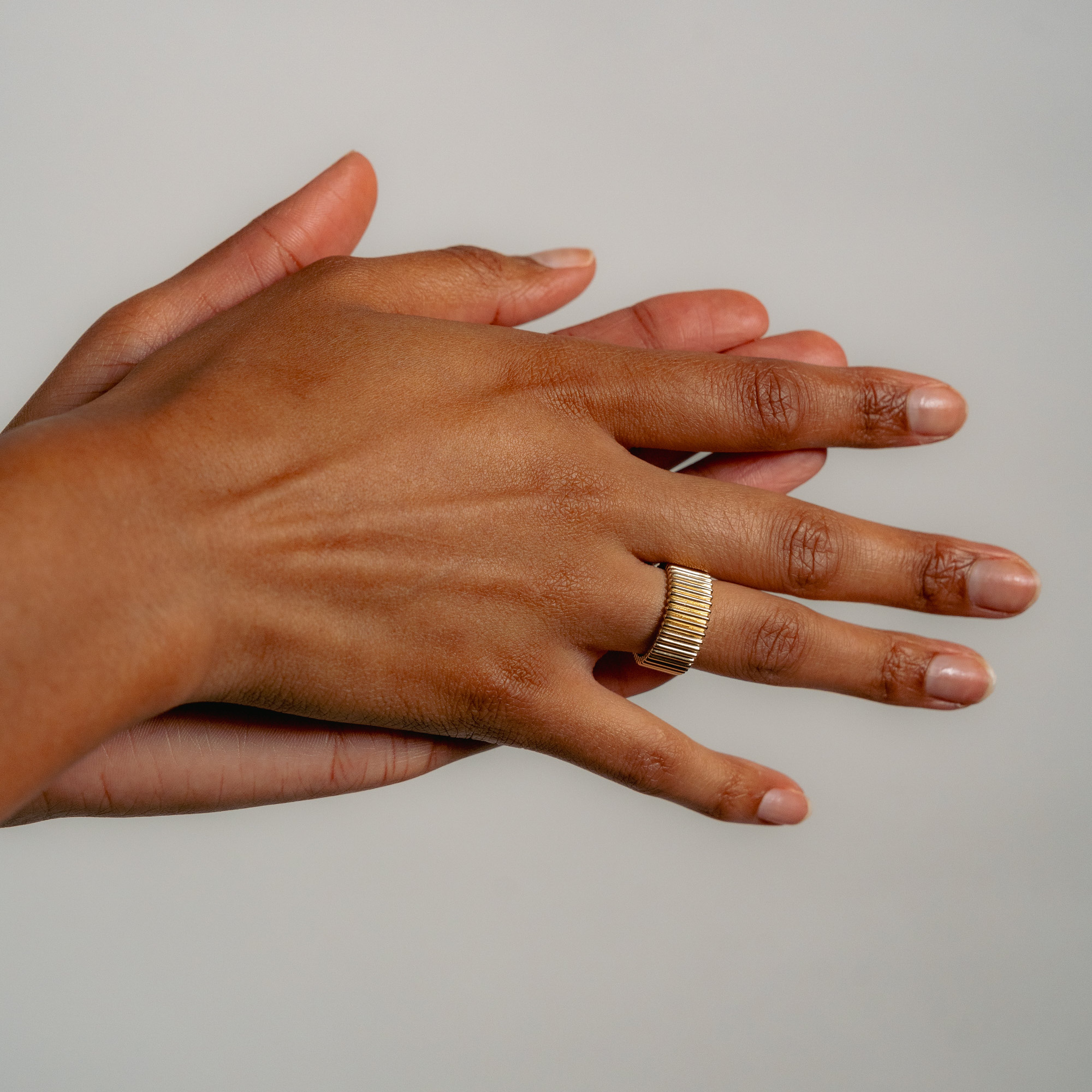 Hands clasped against a gray background, displaying a gold ring with vertical design on one finger, conveying a sense of elegance and calm.