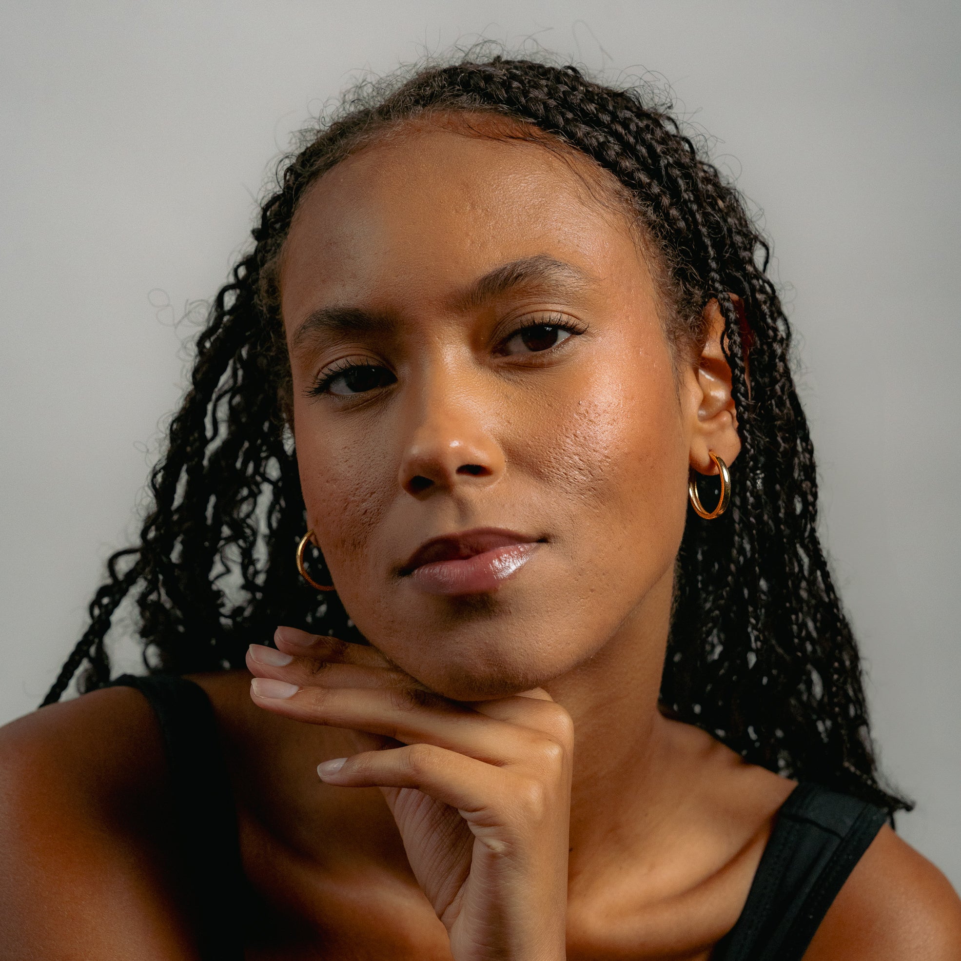 A woman with braided hair models Glasâje’s Gertrude Hoops in 9ct solid gold (23mm diameter), wearing a sleeveless black top and confidently posing with her hand under her chin against a plain background.