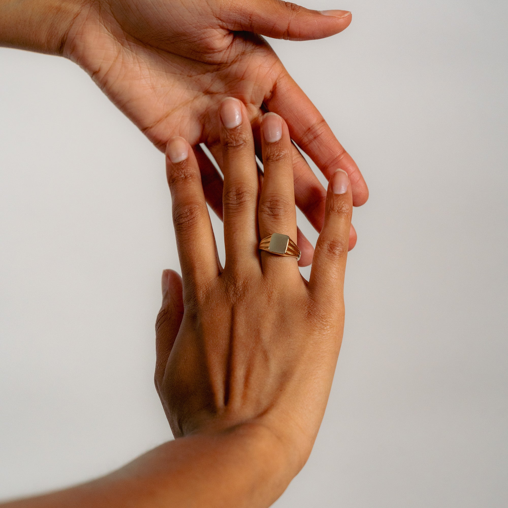 A close-up shows one hand gently touching another, which wears the Glasâje Signet Ring—a solid gold ring featuring a rectangular face with subtle debossed detailing—set against a plain, light-colored background.