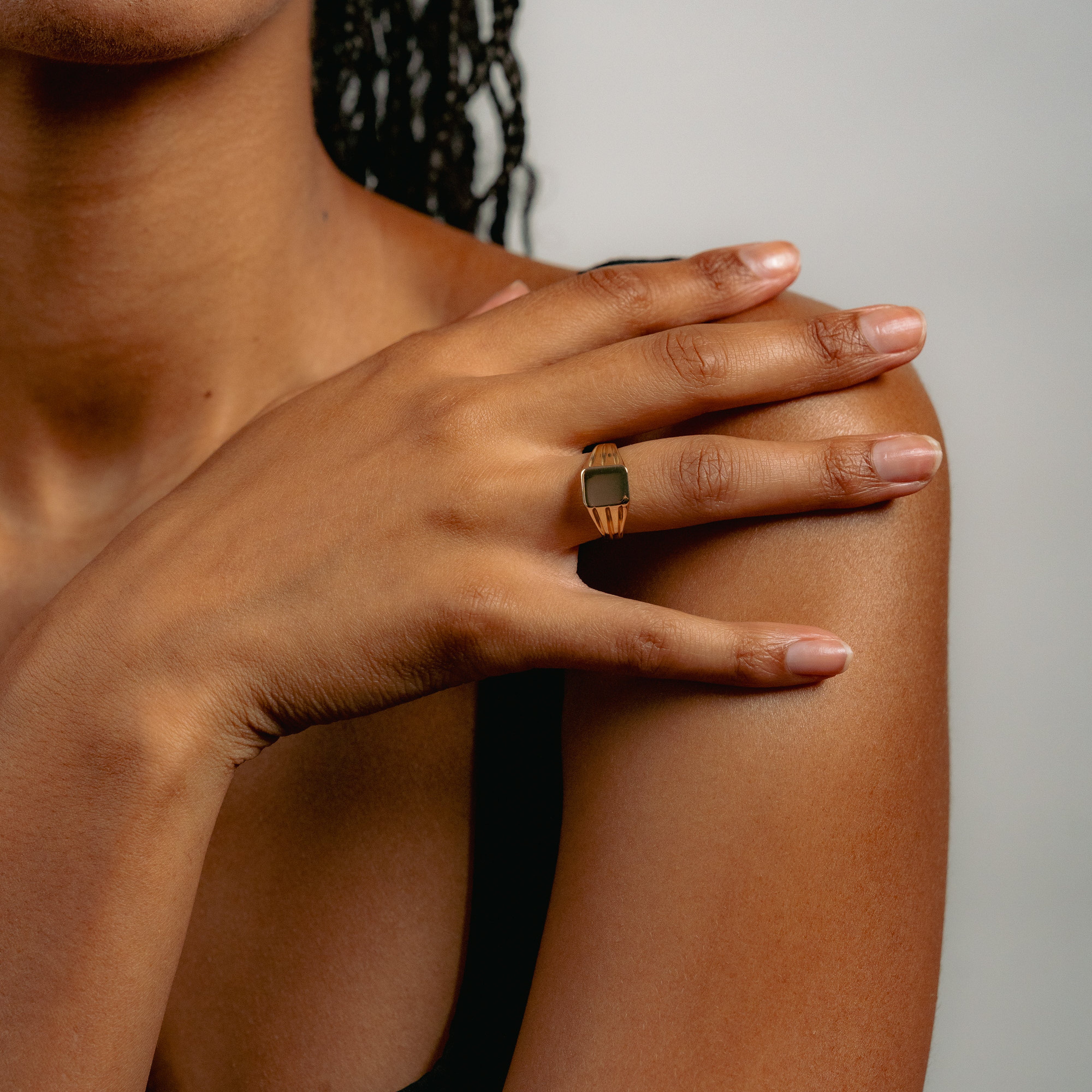A person with medium brown skin showcases the Glasâje Signet Ring, a solid gold piece with a geometric design. Their neatly manicured hand rests on their shoulder, and they wear a black sleeveless top against a softly lit background.